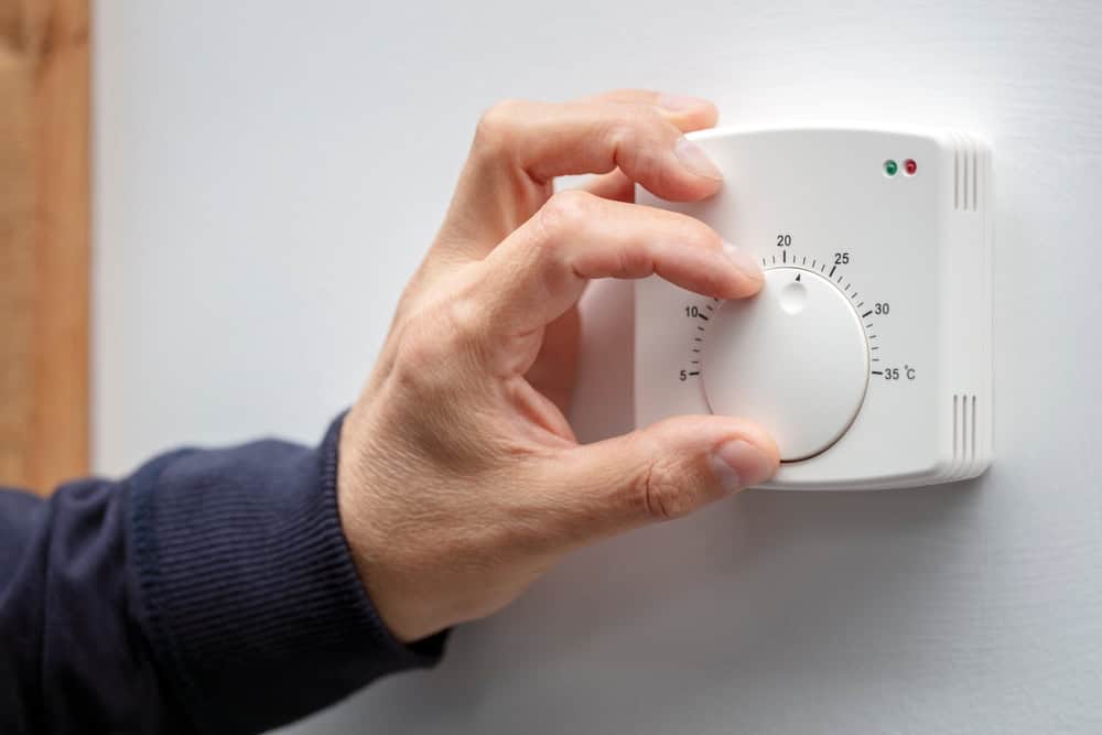 A hand adjusting a round dial thermostat on a wall. The thermostat is set to approximately 22 degrees Celsius, with red and green indicator lights visible above the dial.