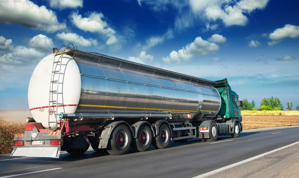 A large green semi-truck with a shiny silver cylindrical tanker trailer drives on a paved highway under a bright blue sky with scattered clouds, passing open fields.